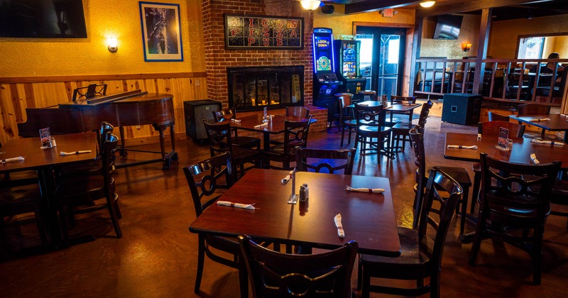 Rustic, warmly lit dining room with wooden tables, a grand piano, brick fireplace, gaming machines, and raised seating