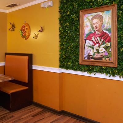 Restaurant corner with booth seating and a framed Frida Kahlo portrait on a greenery wall.