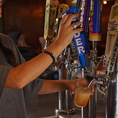 A person pours cider from a blue tap labeled CIDER into a plastic cup.