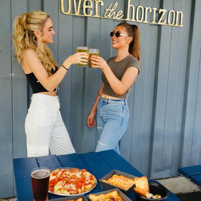 Exterior, two cheery friends stand by a table with food, toasting with beers.