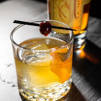 A glass of bourbon cocktail with ice, orange peel, and cherry stick garnish, sits on a wooden table.
