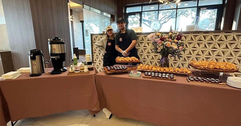 A pair of employees positioned behind a catering table displaying various pastries and coffee