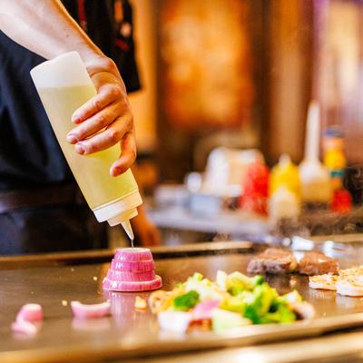 A chef's hand holds a yellow squeeze bottle over stacked onion rings on a grill.