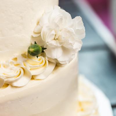 Close-up of a white frosted cake with elegant piping and a white flower accent.