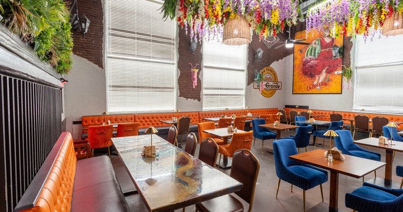 A restaurant interior with orange booth seating, blue and orange velvet chairs, wooden tables, and colorful hanging flowers