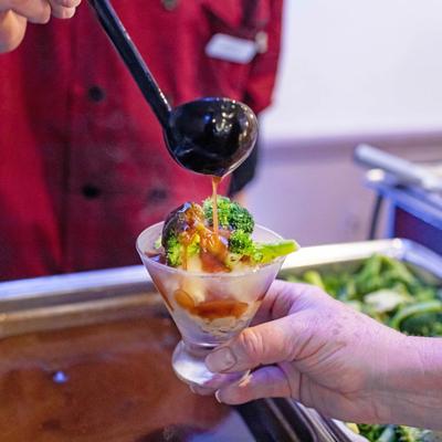 A buffet server pouring brown gravy over mashed potatoes and steamed broccoli in a glass cup.
