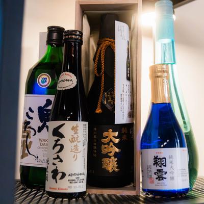 Five Japanese sake bottles stand on a shelf, one in a box.