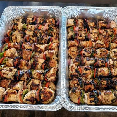 Two aluminum catering trays filled with grilled chicken skewers accompanied by vegetables.