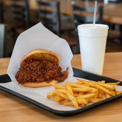 BBQ meat sandwich and fries, served on a wooden table with a cup of beverage.