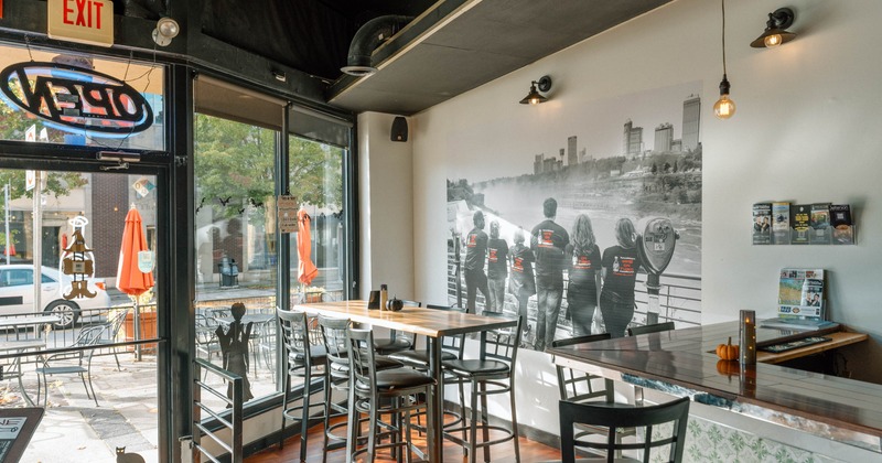 Bright café interior with tall tables, bar stools, large windows, and an outdoor patio visible through the entrance