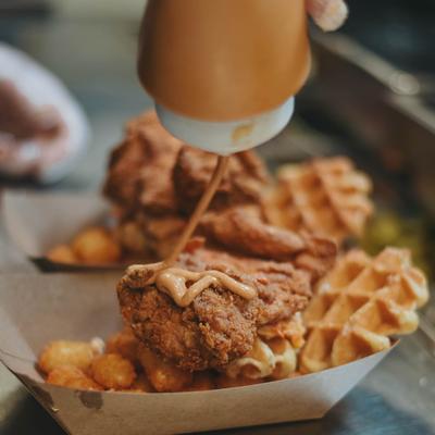 Chef drizzling sauce over fried chicken pieces and waffles served in paper trays.