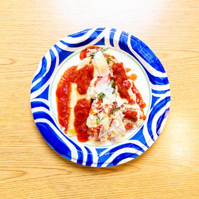 Lasagna served on a blue patterned paper plate, set atop a light wooden table.