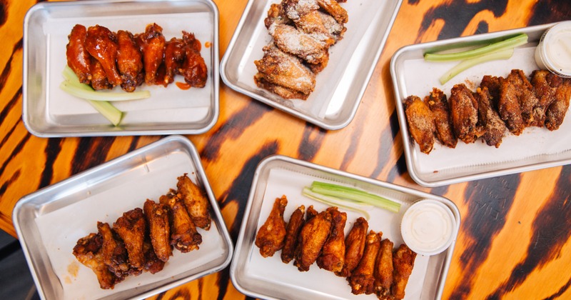 Five trays of assorted chicken wings with celery sticks and ranch on a wooden table
