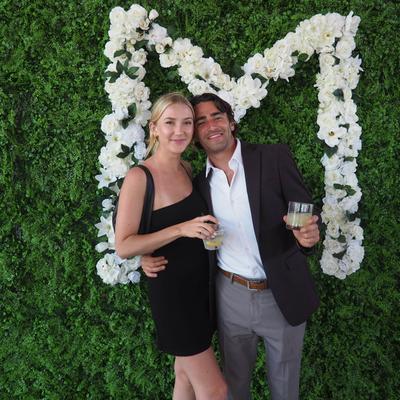 A smiling couple holding their drinks in front of a floral decoration.