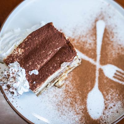 A slice of tiramisu with whipped cream and cocoa powder art on a white plate.