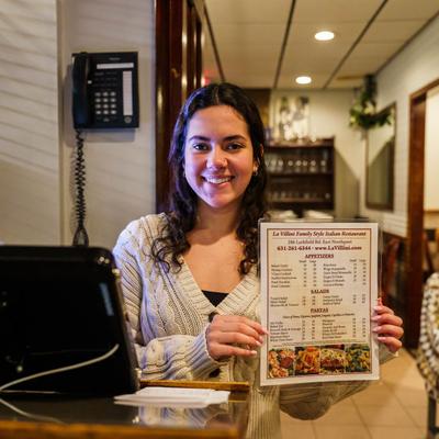 Employee smiling and holding restaurant's menu for the camera behind front desk counter