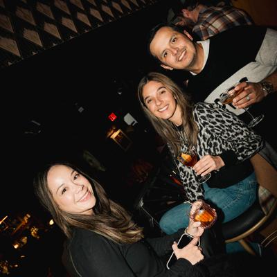 Three people smile while holding drinks at a dimly lit bar setting.