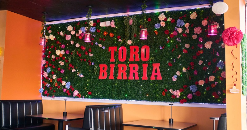 Restaurant interior with a flower-covered wall displaying "TORO BIRRIA" in red letters