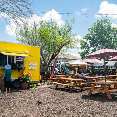 Outdoor scene with a yellow food truck, a person ordering, and picnic tables under umbrellas.