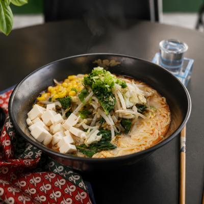 A bowl of ramen with tofu, corn, and greens on a table.