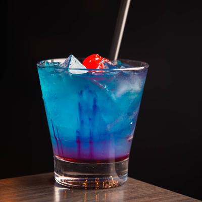 Purple Rain cocktail with a cherry, sits on a wooden table, with a black background.