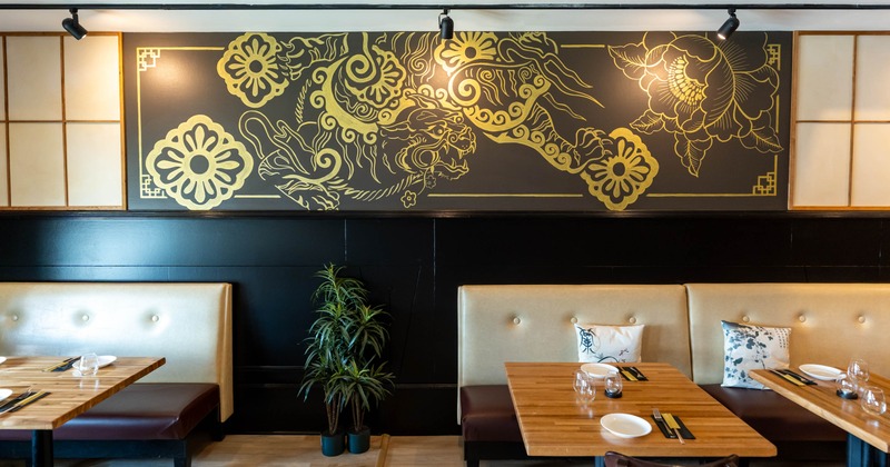 Interior with a gold mural on a black wall, beige seating, wooden tables and a potted plant