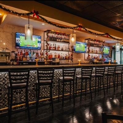 A warmly lit bar adorned, featuring liquor bottles, a line of black bar chairs, and TVs.