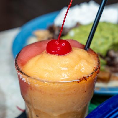 A frozen, layered mango margarita garnished with a maraschino cherry and straw.