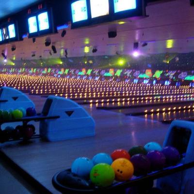 An indoor bowling alley features multiple glowing lanes, balls, and overhead monitors.
