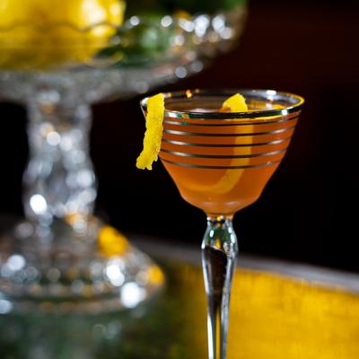 Amber cocktail in a striped glass garnished with long citrus twist on bar.