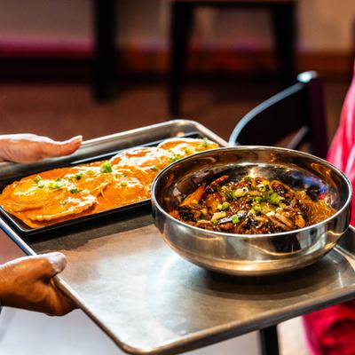 Person holding a metal tray featuring a bowl of beef stir-fried noodles and plate of ravioli.
