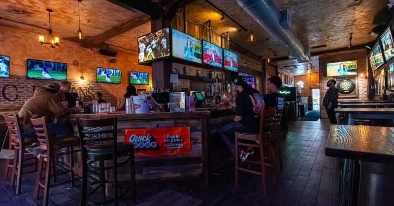 Interior,  corner of bar, stools, guests, screens above drink rack