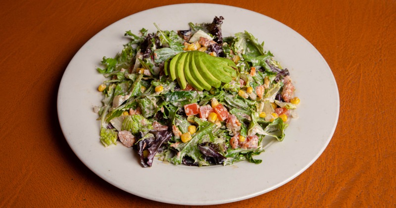 Salad featuring lettuce, chopped tomatoes, and corn, topped with sliced avocado
