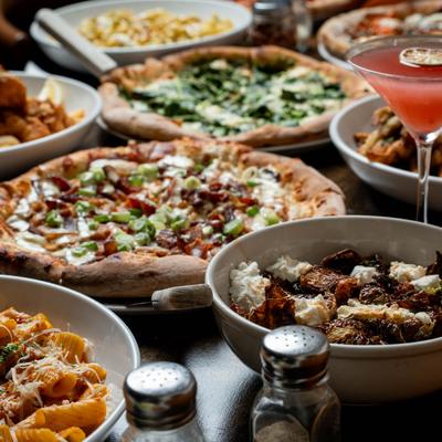 Various pizzas and pasta dishes on a table alongside a pink cocktail.
