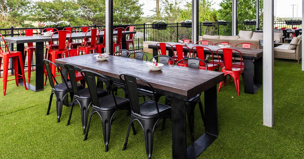 Outdoor dining area with wooden tables and metal chairs on artificial grass.