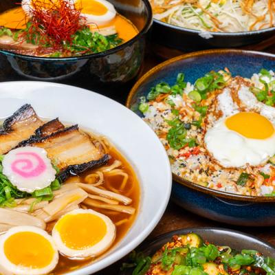 Bowls of ramen with pork and egg beside rice bowls and other dishes on a table.