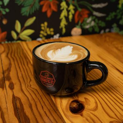 A dark cup of caramel nut latte sits on a wooden table with a floral background.