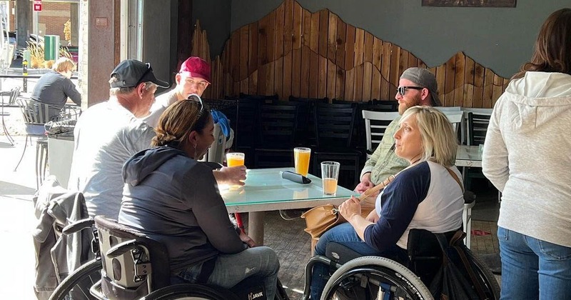A group of three people in wheelchairs sitting around a table eating and laughing at Brewability