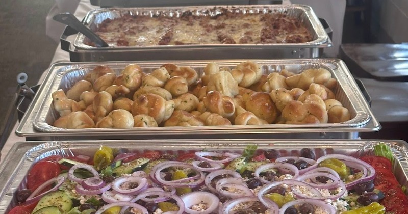 Catering buffet set up with trays of baked pasta, garlic knots and a salad