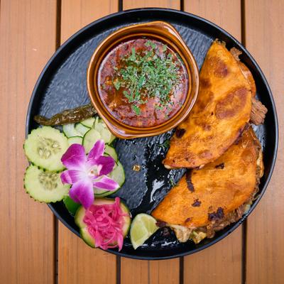Queso birria tacos served with consommé, sliced cucumber, pickled red onions, and lime.