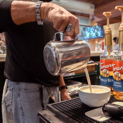Barista pouring milk from a metal pitcher into a white bowl