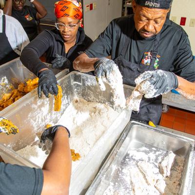 Kitchen staff breading chicken pieces in large containers.