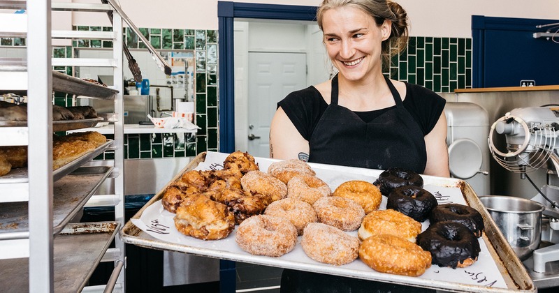 Smiling baker in a black apron holds a tray of assorted donuts