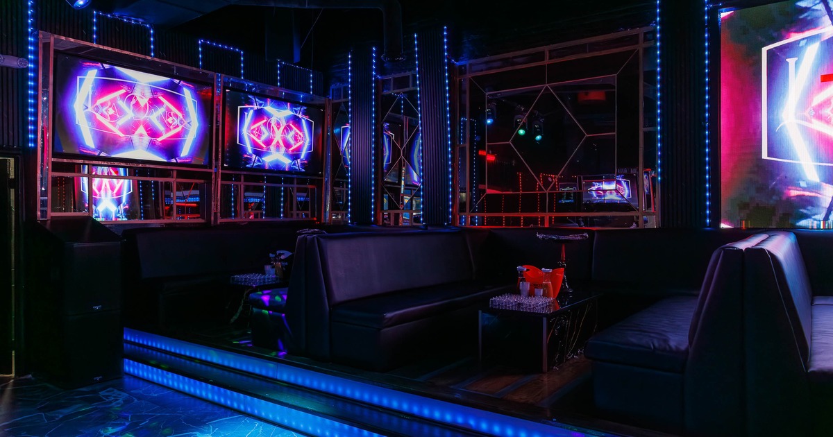 A dark lounge interior with neon-lit screens, black leather couches and blue LED lighting.