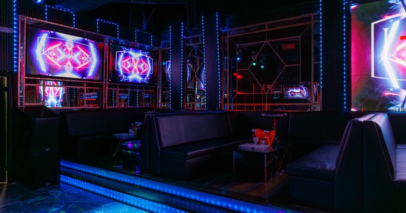 A dark lounge interior with neon-lit screens, black leather couches and blue LED lighting.