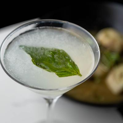 Pale cocktail in a V-shaped martini glass garnished with a basil leaf.