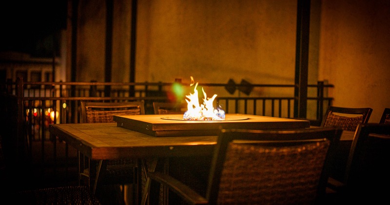 Outdoor table with a central fire pit, surrounded by wicker chairs in a dimly lit setting