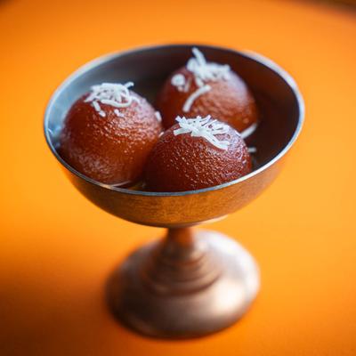 A silver chalice holds three syrupy gulab jamuns garnished with coconut flakes.