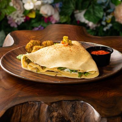 A savory crepe served on a wooden plate with falafel-like side dishes and a small cup of red sauce.