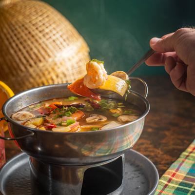 A hand scoops steaming Tom Yum seafood soup from a hot pot.